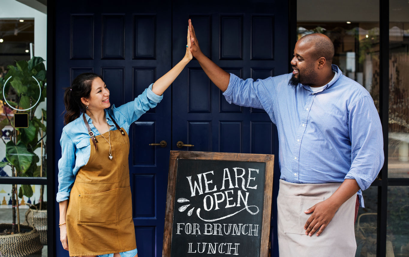 man and woman high five in front of a store font with open sign