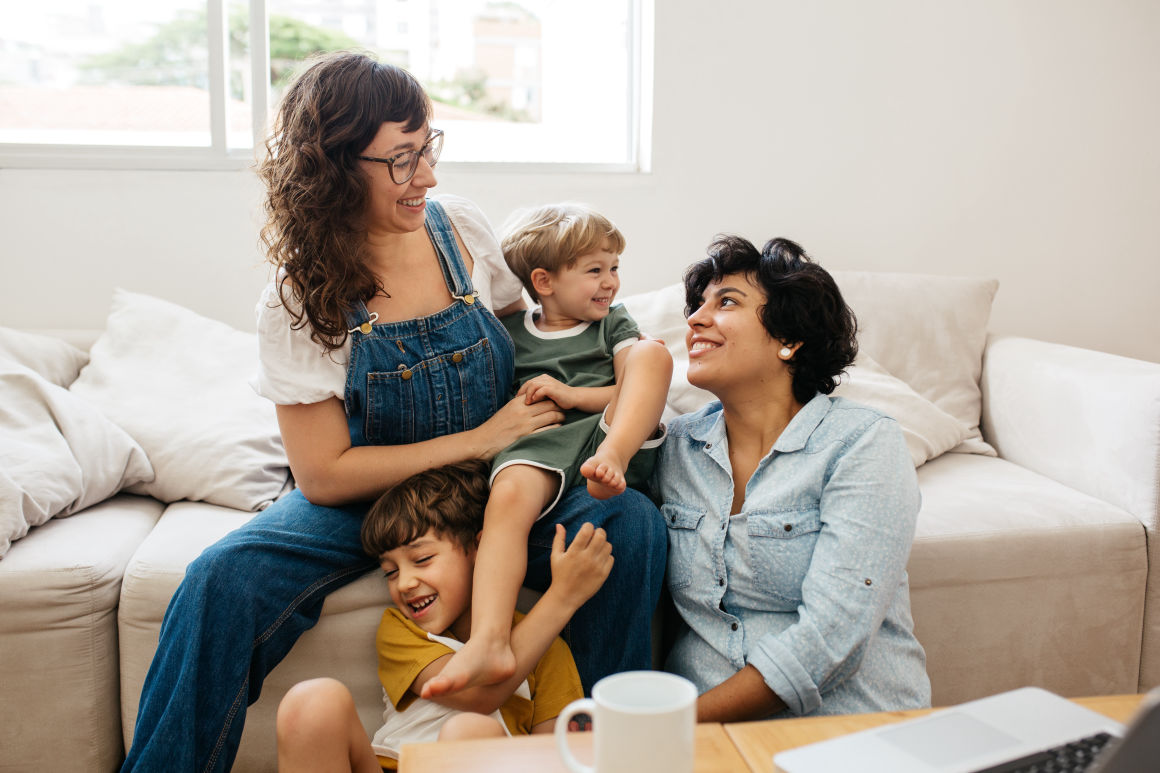 A happy family with two women and two young children laughing together on a sofa in a bright living room