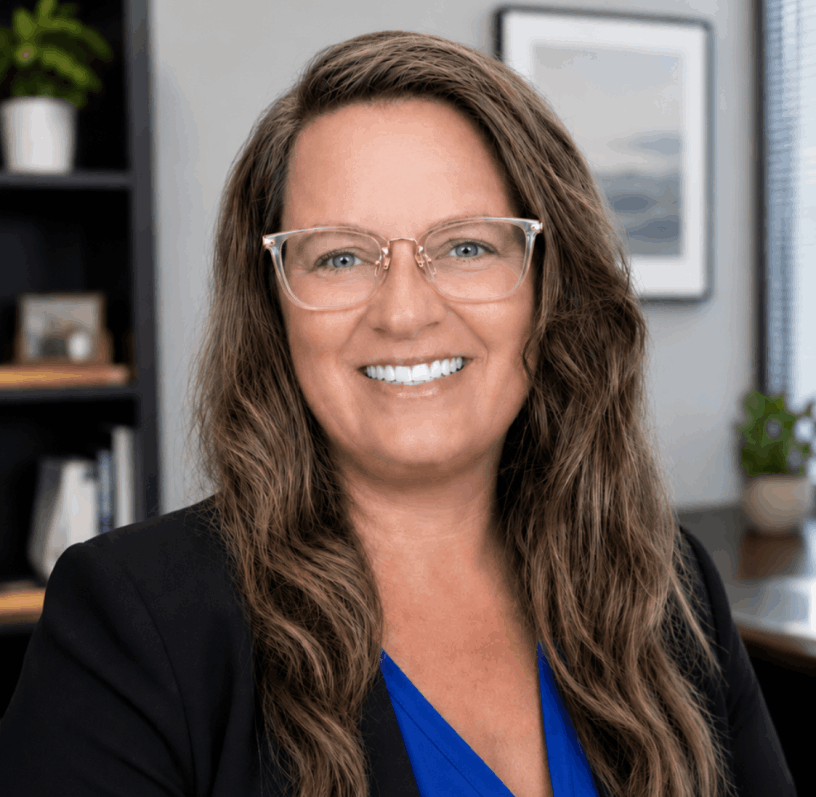Ashley Jean Gorby smiling wearing glasses wearing a black jacket and blue shirt in a office